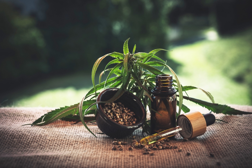 CBD Öl, ein CBD Öl Glas mit einer Hanfpflanze im Hintergrund und ein kleines Schälchien mit Bohnen